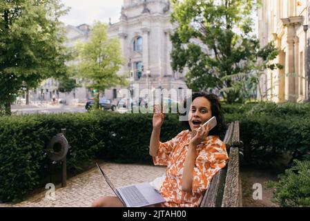 émerveillement jeune indépendant en tenue d'été assis sur banc avec ordinateur portable et parlant sur smartphone dans le parc vert, image de stock Banque D'Images