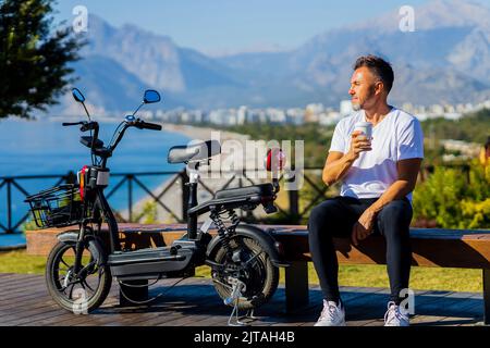 bel homme en t-shirt blanc assis sur le banc à côté du scooter écologique avec belle vue sur les collines et boire un café à emporter Banque D'Images