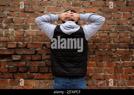 Le gars se tient face à un mur de brique avec ses mains clastées derrière sa tête. Un gars en Jean et un débardeur boursouflé noir avec son dos à l'appareil photo. Arre Banque D'Images