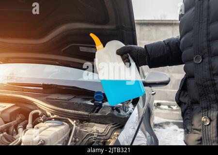 Gros plan de gants conducteur ou mécanicien versant du liquide antigel bleu pour le lavage de pare-brise de voiture d'hiver avec arrosoir bootle Banque D'Images