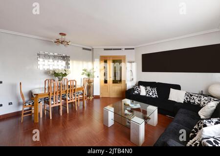 Salle de séjour d'une maison résidentielle avec une table à manger en bois avec chaises assorties, un canapé d'angle couvrant deux murs tapissés de tissu noir et RE Banque D'Images