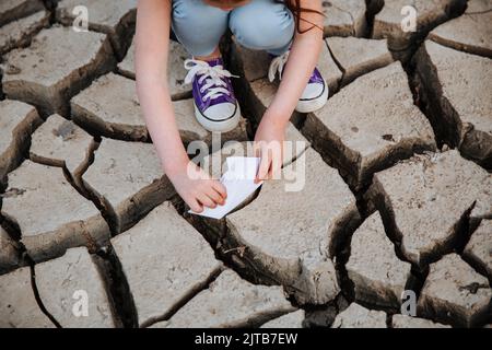 La fille abaisse le bateau en papier sur le sol sec et fissuré. Crise de l'eau et changement climatique concept. Réchauffement de la planète Banque D'Images