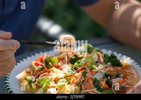 Main mâle tenant des baguettes avec une crevette d'une salade de papaye de fruits de mer en foyer Banque D'Images