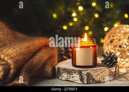 Carte d'hiver confortable avec bougie parfumée dans un pot en verre sombre avec maquette d'étiquette vide et pull en laine sur des lumières de bokeh floues sur l'arrière-plan. Banque D'Images