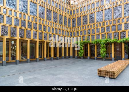 Le Stadhuiskwartier à Deventer, l'une des neuf villes hanséatiques historiques sur la rivière IJssel, province d'Overijssel, Pays-Bas, est l'extension du hôtel de ville achevée en 2016 par les architectes Neutelings Riedijk. Sa façade en verre encadrée de jaune porte l'œuvre d'art "Deventer Raamwerk" (Cadre de Deventer) de l'artiste Loes ten Anscher : 2 264 empreintes digitales de résidents de Deventer moulées en aluminium. Vu depuis le coin opposé de la cour, la vue s'ouvre sur l'entrée vitrée "Stadhuis" avec de la verdure grimpante et un banc en bois au premier plan. Banque D'Images
