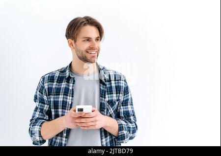 Positif beau style caucasien gars, tenant le smartphone dans la main, chat en ligne, la navigation sur Internet, regardant le côté, penser, rêver, debout sur un arrière-plan blanc isolé, sourire Banque D'Images