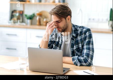 Frustré triste homme caucasien, yeux fermés, massages son nez de pont, assis à son lieu de travail à la maison bureau dans la cuisine, prendre une pause. Se sentir épuisé et fatigué pendant de longues heures de travail dans l'ordinateur portable Banque D'Images