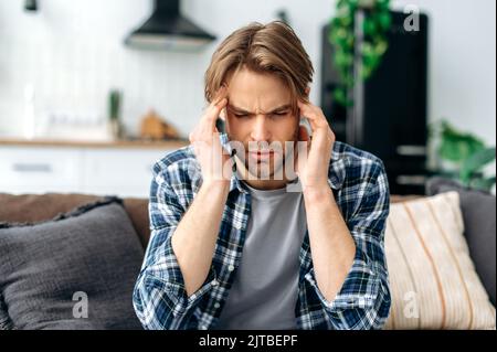 Gros plan de l'homme exténué caucasien, assis sur un canapé dans la salle de séjour, massages de ses temples, fermé ses yeux, se sent mal, souffrant de migraine, fatigue, besoin de traitement, repos. Maux de tête, spasme Banque D'Images