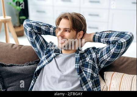 Gros plan d'un caucasien calme et détendu en chemise décontractée, reposant sur un canapé confortable dans un salon confortable, jetant ses mains derrière sa tête, rêvant de vacances, se reposant du travail, souriant Banque D'Images