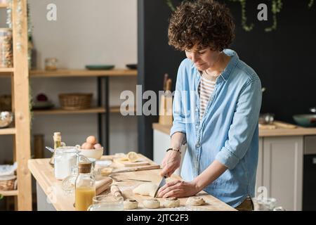 Jeune femme coupant de la pâte pour de petits morceaux pour cuire des petits pains faits maison à table dans la cuisine Banque D'Images