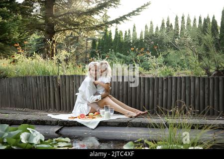 Portrait de famille assis sur un plat avec des fruits, pot de lait sur le bord de l'étang. Jeune femme embrassant petite fille fille fille. Banque D'Images