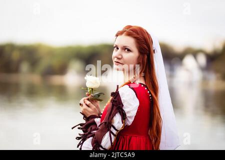 Gros plan d'une fille mince à cheveux rouges dans une robe rouge à la mode vénitienne du 15th siècle avec une rose blanche sur fond d'eau Banque D'Images