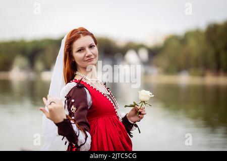 Fille mince à cheveux rouges vêtie d'une robe rouge à la mode vénitienne du 15th siècle avec une rose blanche sur fond d'eau Banque D'Images