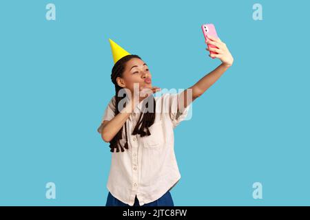 Femme romantique en cône diffusant en direct, envoyant un baiser aérien à des abonnés, enregistrant une vidéo pour le blog pendant l'anniversaire de célébration, portant une chemise blanche. Studio d'intérieur isolé sur fond bleu Banque D'Images