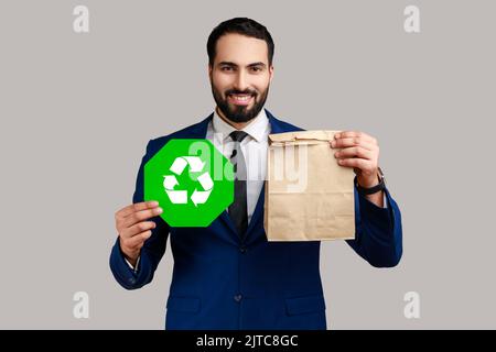 Portrait d'un homme barbu souriant tenant une affiche de recyclage verte à la main et un emballage en papier, pensant vert, portant un costume de style officiel. Prise de vue en studio isolée sur fond gris. Banque D'Images