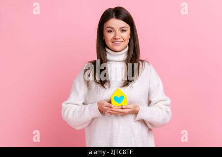 Belle femme a le rêve et la tenue de papier petite maison, la planification de la nouvelle maison à acheter, regarde l'appareil photo, portant blanc style décontracté pull. Studio d'intérieur isolé sur fond rose. Banque D'Images