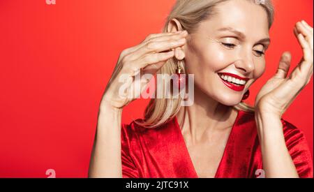 Magnifique femme mûre aux lèvres rouges, boucles d'oreilles et robe de vacances, dansant nad rire, debout sur fond de studio Banque D'Images