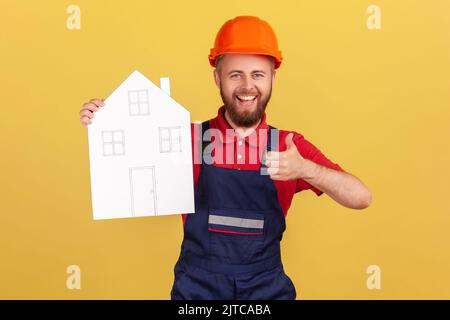 Portrait de l'homme de construction tenant la maison de papier, offrant de l'aide dans la réinstallation, recommander des services de réparation à domicile, montrant le pouce vers le haut, portant des combinaisons et un chapeau. Studio d'intérieur isolé sur fond jaune. Banque D'Images