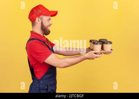 Portrait de côté d'un sympathique messager heureux dans des combinaisons bleues donnant du café, donnant des boissons dans des tasses jetables, exprimant des émotions positives. Studio d'intérieur isolé sur fond jaune. Banque D'Images