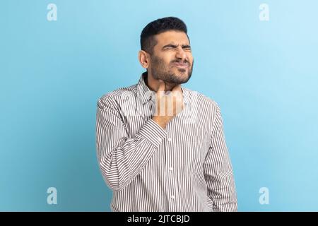Douleur à la gorge. Portrait d'un homme d'affaires malade malheureux touchant son cou, souffrant de maux de gorge, d'infection virale ou de symptômes de grippe, portant une chemise rayée. Studio d'intérieur isolé sur fond bleu. Banque D'Images