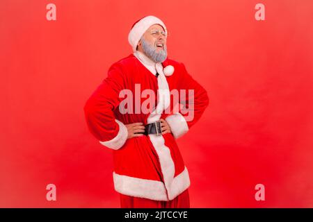 Portrait d'un homme âgé avec une barbe grise portant le costume du père noël debout et gardant les mains sur le ventre, souffrant de douleur, de gastrite ou de constipation. Studio d'intérieur isolé sur fond rouge. Banque D'Images