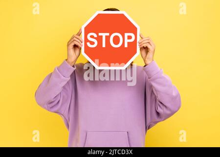 Femme inconnue couvrant le visage avec le symbole Stop, personne anonyme portant le panneau de signalisation rouge, avertissement sur les règles de sécurité routière, port de capuchon violet. Studio d'intérieur isolé sur fond jaune. Banque D'Images