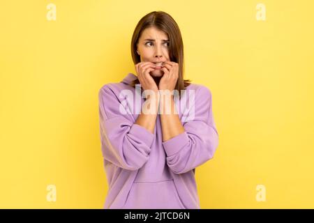 Problèmes et soucis. Portrait de la femme nerveuse piquant des ongles, terrifiée au sujet des problèmes, la phobie de souffrance, le désordre d'anxiété, portant le capuchon pourpre. Studio d'intérieur isolé sur fond jaune. Banque D'Images