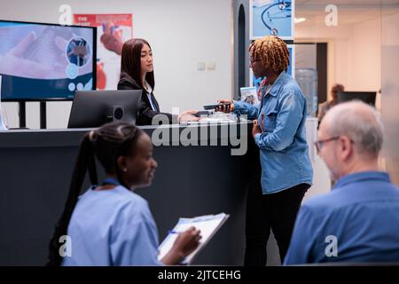 Femme afro-américaine prenant rendez-vous avec transaction par carte de crédit à la réception de l'hôpital. Paiement électronique après avoir effectué une consultation de visite de contrôle dans un centre de santé. Banque D'Images