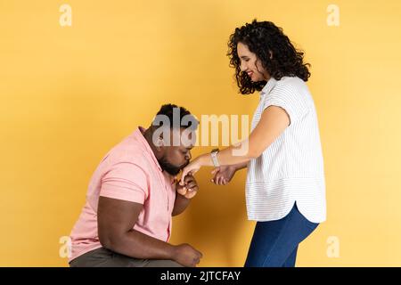 Portrait de jeune couple romantique en vêtements décontractés se posant ensemble, homme debout sur son genou et embrassant sa main de femme, exprimant l'amour. Studio d'intérieur isolé sur fond jaune. Banque D'Images