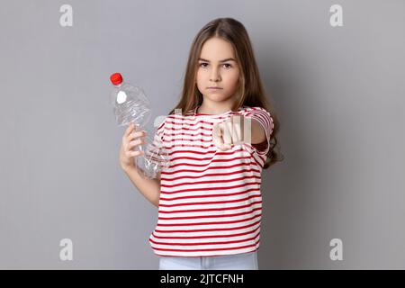 Portrait d'une petite fille sérieuse portant un T-shirt rayé tenant une bouteille en plastique vide, regardant et pointant le doigt vers l'appareil photo. Prise de vue en studio isolée sur fond gris. Banque D'Images