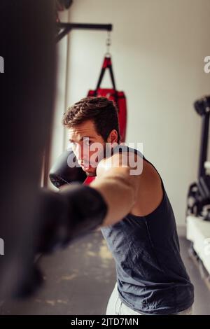 Boxeur musclé frappant un sac de poinçonnage avec un poing ganté. Jeune homme sportif pratiquant ses techniques de poinçonnage pendant une session d'entraînement à la gy Banque D'Images