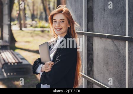 Femme d'affaires gaie avec un visage rousseté et des cheveux rouges attendant son partenaire dans le parc tenant un ordinateur portable. Belle fille attrayante. Jour ensoleillé. Magnifique Banque D'Images