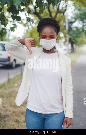 Jeune femme afro-américaine portant un masque médical sur montrer le pouce vers le bas geste, marchant dans la rue. Personnel de santé concept de médecine de santé. Banque D'Images