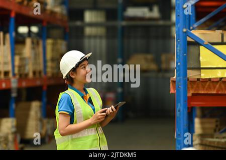 Jeune femme travaillant en entrepôt dans des casques et des vestes réfléchissantes utilisant une tablette numérique et des boîtes de comptage sur la tablette Banque D'Images