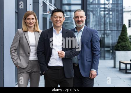 Équipe d'affaires diversifiée réussie, trois employés souriant et regardant la caméra, équipe de rêve avec un patron asiatique à l'extérieur du bâtiment de bureau, des collègues dans des costumes d'affaires Banque D'Images
