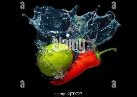 Le piment rouge chaud et la chaux toute fraîche sont tombés sous l'eau avec des éclaboussures isolées sur fond noir. Banque D'Images