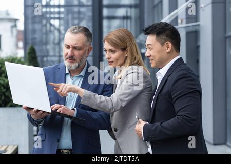 Équipe d'affaires réussie, trois collègues homme d'affaires et femme d'affaires à l'extérieur du bâtiment de bureau discutant des plans et de la gestion actuels, regardant l'écran d'ordinateur portable, discuter et conseiller Banque D'Images