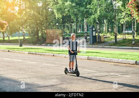 Un écolier fait du scooter électrique sur la route dans un parc d'été vide. Un enfant en t-shirt noir et en short denim se déplace sur un scooter le long d'une route asphaltée marquée Banque D'Images