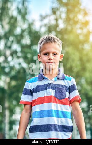 Joli garçon blond avec une coiffure courte dans des vêtements élégants et décontractés posant pour un appareil photo dans le parc vert ensoleillé de la ville d'été Banque D'Images