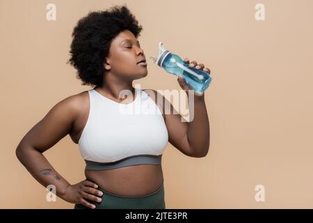 femme afro-américaine de taille plus tenant une bouteille de sport avec de l'eau et debout avec la main sur la hanche isolée sur beige, image de stock Banque D'Images