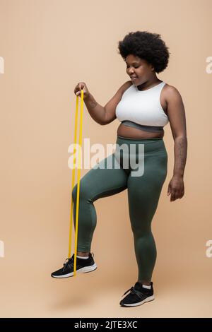 pleine longueur de happy african american taille plus femme en vêtement de sport s'exerçant avec bande de résistance sur beige, image de stock Banque D'Images