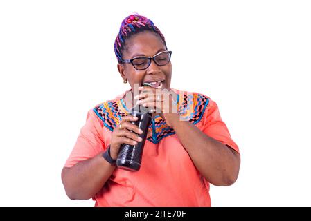une femme africaine en verre debout sur un fond blanc essayant d'ouvrir une bouteille de bière en utilisant ses dents. Banque D'Images