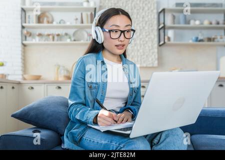 Jeune femme asiatique qui étudie à distance à la maison, étudiante qui écoute des cours en ligne, prend des notes, s'asseyant sur un canapé dans le salon à l'aide d'un casque et d'un ordinateur portable Banque D'Images