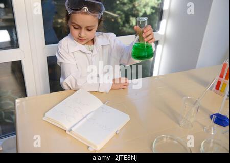 Une petite fille charmante chimiste secoue une fiole avec une solution verte tout en lisant la formule chimique dans le manuel de chimie Banque D'Images