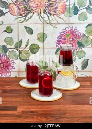 Thé glacé à la framboise dans une théière en verre spéciale, avec deux verres à thé glacé et des pailles borosilicates, sur une table en noyer et des carreaux de céramique en arrière-plan. Banque D'Images