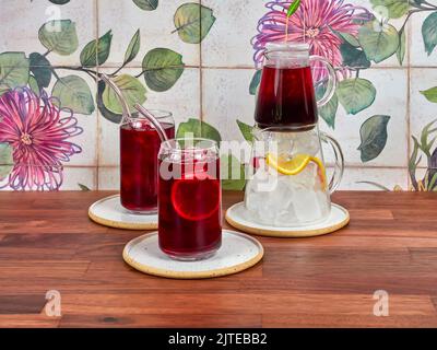 Thé glacé à la framboise dans une théière en verre spéciale, avec deux verres à thé glacé et des pailles borosilicates, sur une table en noyer et des carreaux de céramique en arrière-plan. Banque D'Images