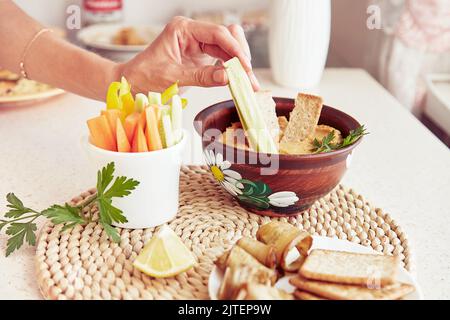 Femme trempez le concombre frais dans un houmous naturel fait maison. Biscuits, persil et légumes frais sur la table. En-cas sains végétariens rustiques en gros plan Banque D'Images
