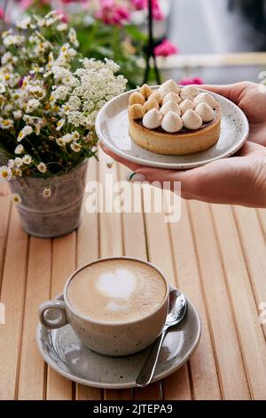 Une femme tient une tarte aux noisettes et au cappuccino sur une table en bois à l'extérieur de la terrasse par temps d'automne froid. Petit déjeuner esthétique. Banque D'Images
