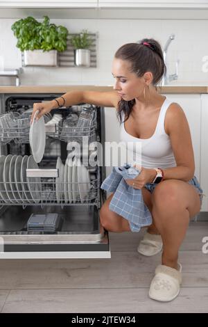 Femme avec un chiffon dans sa main prend la vaisselle hors du lave-vaisselle dans la cuisine. Banque D'Images
