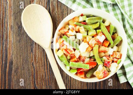 Un mélange de légumes surgelés assortis dans un bol blanc prêt à cuire sur une table en bois brun. Flat lay, vue de dessus avec espace de copie pour le texte Banque D'Images